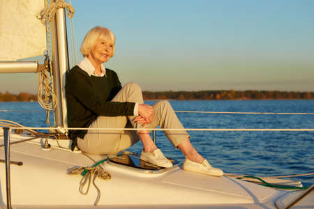 Calm and relax. Beautiful senior woman sitting on the side of sailboat or yacht deck floating in the calm blue sea at sunset, enjoying amazing view and smilingの写真素材