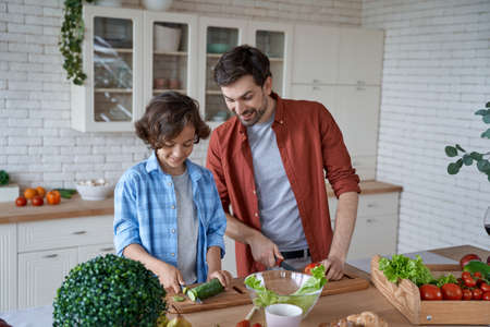 Cooking dinner. Young father teaching his son how to cut vegetables, preparing a salad while standing in the modern kitchen at homeの写真素材