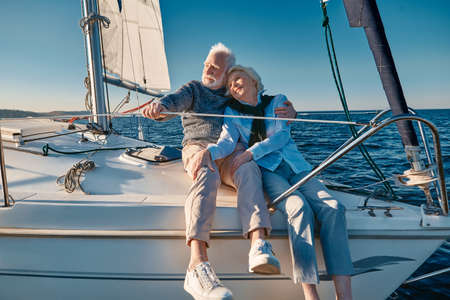 Enjoying sailing. Happy lovely senior family couple hugging and relaxing on a sail boat or yacht deck floating in a calm blue sea, looking at the horizonの写真素材