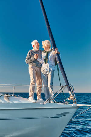 Vacation. Full length of happy senior couple in love holding hands and looking at each other while standing on the side of sailboat or yacht deck floating in seaの写真素材
