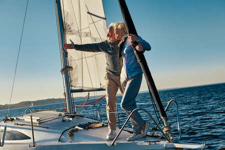 Elegant senior man pointing at the horizon while standing with his wife on the side of a sail boat or yacht deck floating in a calm blue sea, enjoying amazing viewの写真素材