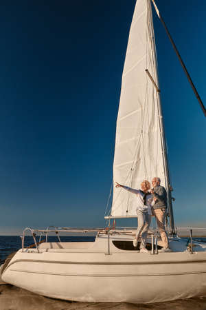 Luxury travel. Full length of happy mature couple standing on the side of sail boat or yacht deck floating in sea, pointing at the horizon, enjoying amazing sunsetの写真素材
