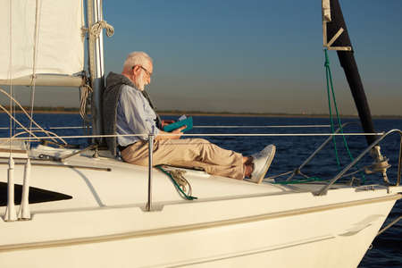 Senior relaxed man reading book while sitting on the side of sailboat or yacht deck floating in the calm blue sea on a sunny dayの写真素材