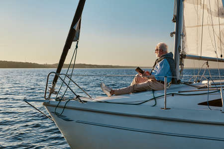 Luxury lifestyle. Side view of a relaxed senior man sitting on the side of sailboat or yacht floating in the calm blue sea at sunset and enjoying amazing view, using digital tabletの写真素材
