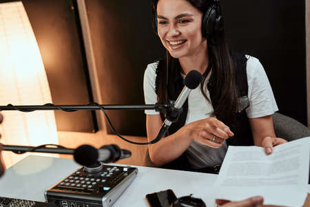 Portait of happy female radio host smiling and giving a script paper to her male colleague while moderating a live show in studioの写真素材