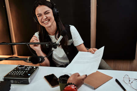 Portait of happy female radio host smiling, receiving a script paper from her male colleague while moderating a live show in studioの写真素材