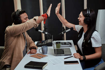 Portait of two cheerful radio hosts, young man and woman giving high five while talking, moderating a live show in studioの写真素材