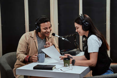 Portait of two radio hosts, man and woman talking with each other, reading a script while getting ready for a live show in studioの写真素材