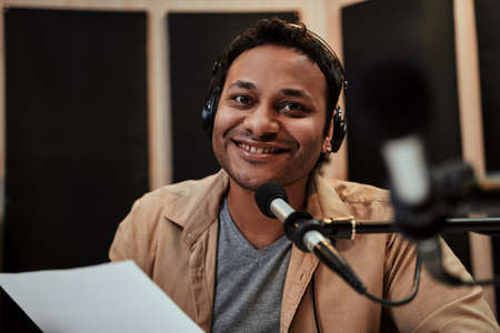 Portrait of happy young male radio host in headphones smiling at camera while talking, broadcasting in studioの写真素材