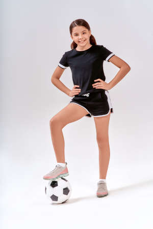 Full-length shot of a cute teenage girl smiling at camera while exercising with soccer ball, isolated over grey background in studioの写真素材