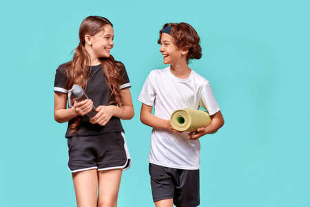 Ready for training. Two teenagers, boy and girl with yoga mat and bottle of water talking and smiling while standing isolated over blue backgroundの写真素材