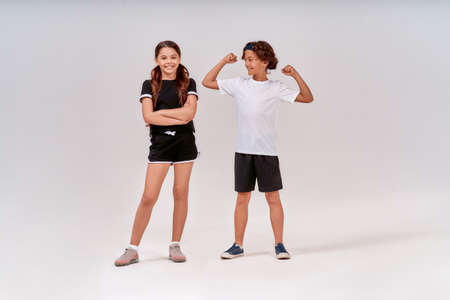 Two teenagers having workout. Happy boy showing his biceps to a cute smiling girl, standing isolated over grey background in studioの写真素材