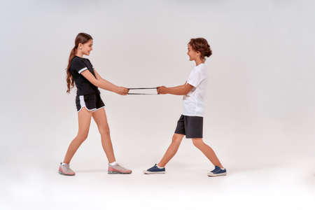 Full-length shot of happy teenage boy and girl having fun while exercising with resistance band, standing isolated over grey background