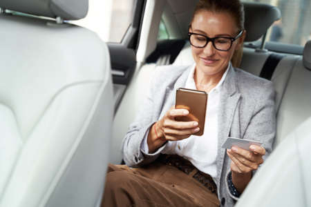 Happy successful business woman wearing eyeglasses using her smartphone and credit card to buy something while sitting on back seat in the carの写真素材