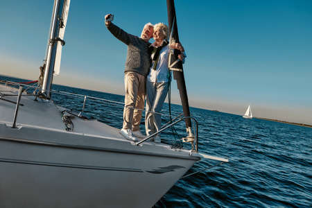 Capturing bright moments. Beautiful senior couple making selfie on smartphone while standing on the side of sailboat or yacht deck floating in seaの写真素材