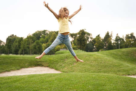 Happy childhood concept. Cute little girl in casual clothes jumping and having fun while visiting park on a sunny summer day, she is smiling and feeling so excitedの写真素材