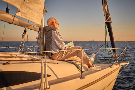 Sailing man. Senior man sitting on the side of his sailboat or yacht floating in the sea, reading book and relaxing, enjoying sunsetの写真素材