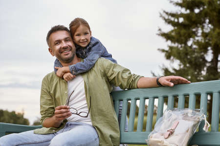 I love my daddy. Portrait of happy father and little daughter sitting on the wooden bench in park, hugging, looking at camera and smilingの写真素材