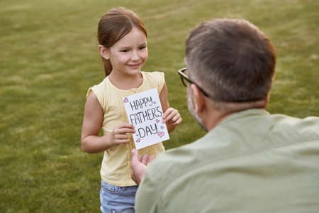 Father and his cute little daughter celebrating Happy Fathers Day in park on a warm day. Small girl giving her father handmade postcard and smilingの写真素材
