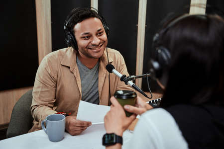 Portrait of young male radio host going live on air, talking with female guest, holding a script paper while sitting in studioの写真素材