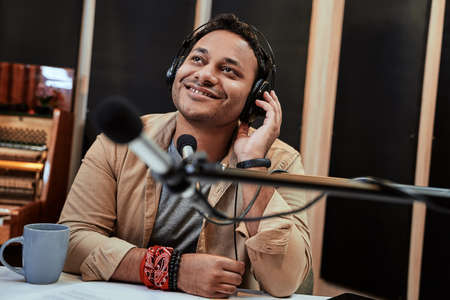 Happy young male radio host looking aside while broadcasting in studio, using microphone and...