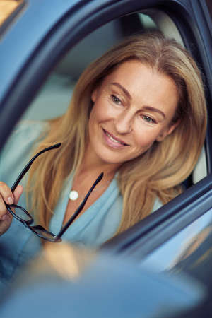 Happy mature business woman sitting behind steering wheel of her modern car, holding eyeglasses and...