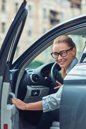 Great day. Beautiful and happy middle aged business woman in classic wear getting out of her modern car, she is arrived at work in the morningの写真素材