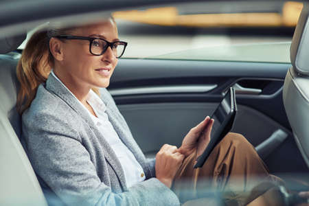 Side view of happy beautiful business woman sitting on back seat in the car and working online, she is digital tabletの写真素材