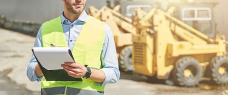 Young civil engineer wearing helmet writing report while standing at construction siteの写真素材