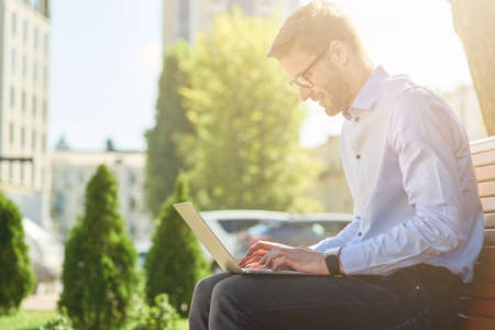 Side view of a handsome male freelancer or businessman working online on laptop while sitting on the bench outdoors on a sunny dayの写真素材