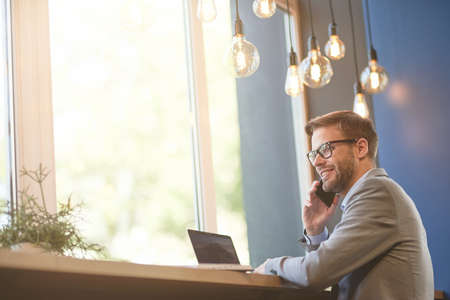 Good news. Side view of a happy young businessman talking by phone and smiling while working remotely in cafeの写真素材