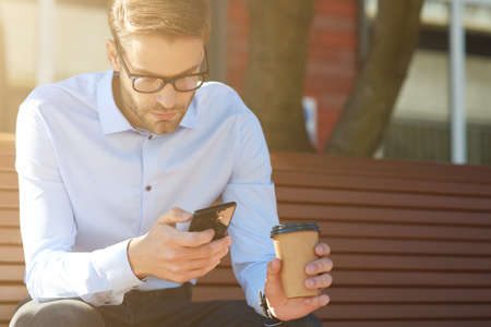 Business message. Focused businessman using smartphone and drinking coffee while sitting on the bench outdoorsの写真素材