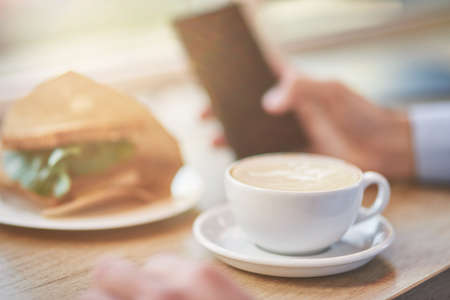 Energy drink. Cropped shot of a man using smartphone while having breakfast in cafe. Cup of coffee and fresh sandwich on wooden tableの写真素材