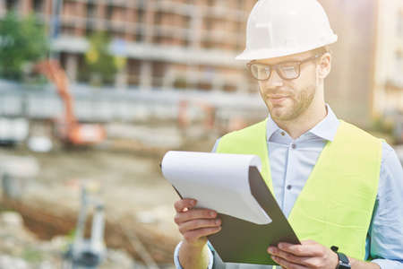 Controlling the project. Young focused civil engineer or construction supervisor wearing helmet reading report while inspecting construction siteの写真素材