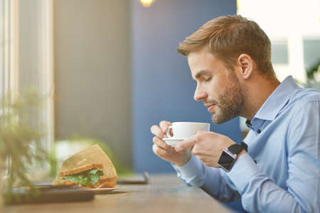 Breakfast time. Side view of a young businessman drinking coffee and going to eat fresh sandwich while sitting in cafeの写真素材