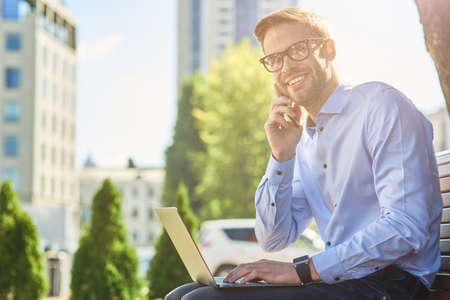 Good news. Young happy businessman talking by phone and and smiling while sitting on the bench outdoors and working online, using laptopの写真素材