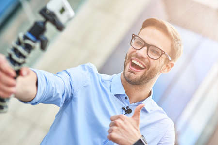 Young happy man, male blogger wearing blue shirt holding a gimbal with smartphone and recording video for his blog outdoors, smiling at camera and showing thumb upの写真素材