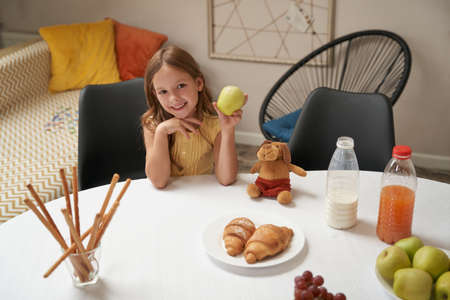 Happy caucasian girl sitting at the table at home, holding green apple and smiling at camera, having breakfast at homeの写真素材