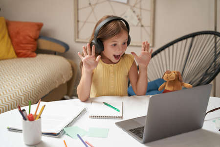 Having video call with teacher. Happy little caucasian girl in headphones studying online, waving and smiling. School during coronavirus pandemicの写真素材