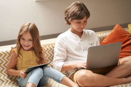 Happy little boy and girl, brother and sister sitting on sofa at home, using laptop and digital tablet and watching cartoons or playing games onlineの写真素材