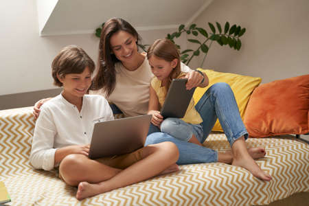 Watching cartoons online. Young beautiful caucasian family, mother and two cute little kids relaxing on sofa together and looking at laptop screenの写真素材
