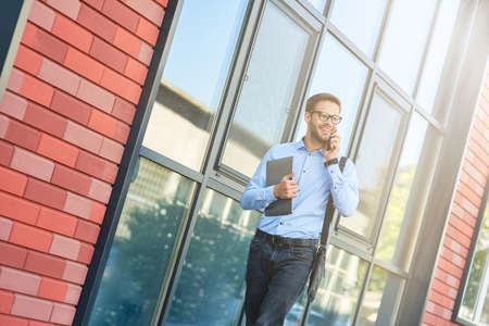 Young happy caucasian businessman wearing blue shirt and eyeglasses holding his laptop, talking by phone and smiling while walking outdoorsの写真素材