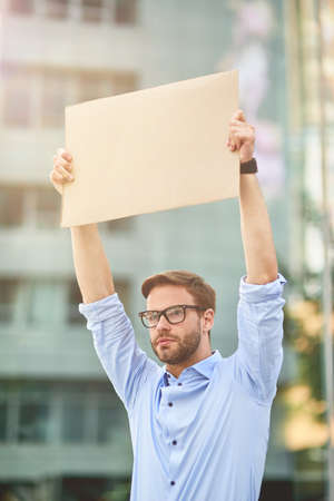 Portrait of a young male activist wearing blue shirt and eyeglasses holding empty sign board while standing outdoors, vertical shotの写真素材