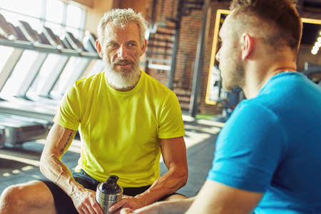 Resting after workout. Focused middle aged man in sportswear holding bottle of water and talking with fitness instructor at gym or sport clubの写真素材