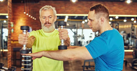 Focused mature caucasian man in sportswear doing weight exercises with assistance of a young professional personal trainerの写真素材
