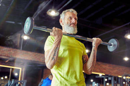 Weight workout. Strong mature man in sportswear lifting weighted bar or barbell at gymの写真素材