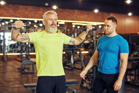 Training body in any age. Focused mature caucasian man in sportswear exercising with dumbbells under supervision of a young professional personal trainer at gymの写真素材