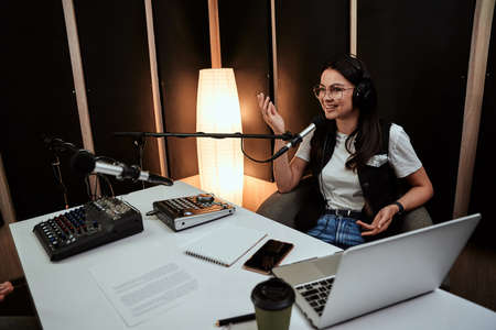Portait of attractive young female radio host looking cheerful while speaking in microphone, moderating a live showの写真素材