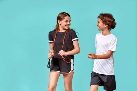 Two happy teenagers, boy and girl with jumping rope talking and smiling while standing isolated over blue background, resting after workoutの写真素材