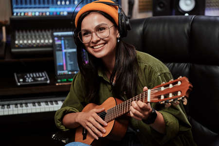 Portrait of beautiful young brunette, female artist smiling aside, playing ukulele while sitting in recording studioの写真素材